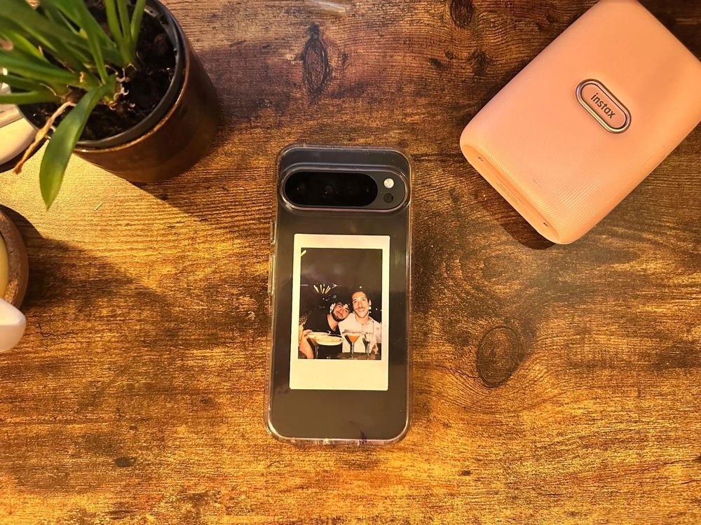 an instax mini print of two friends tucked inside a clear phone case, on a wooden desk beside a pink instax printer